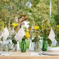Einfache Tischdeko Sommer: Gelbe Blumen in grünen Flaschen, Weingläser und kleine Fähnchen mit der Aufschrift "Enjoy every moment" auf einem Holztisch im Freien.